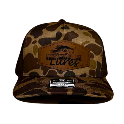 Front view of the Brook Duck Camo Brown Thermocline Lures hat with a brown leather patch logo on camo fabric and adjustable fit.