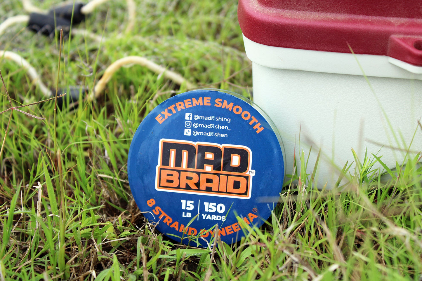 Mad Braid braided fishing line spool placed on grass beside a tackle box, emphasizing its rugged durability and smooth casting ability.