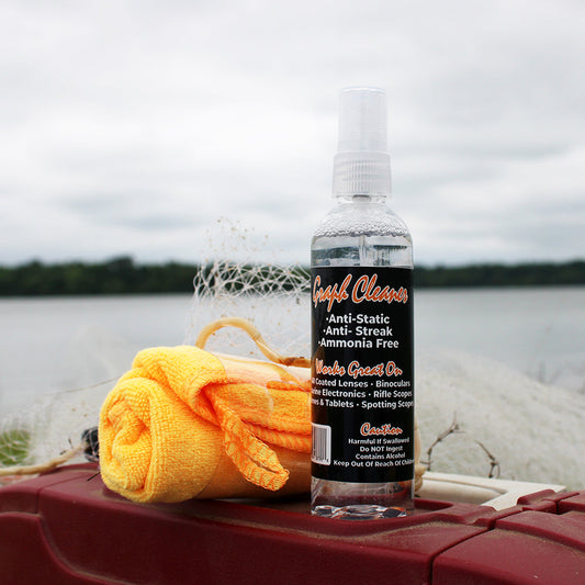 MadFishen Graph Cleaner bottle placed on a tackle box near a lake, with a microfiber cloth. Anti-static, streak-free, and ammonia-free, perfect for cleaning fish finders, marine electronics, and coated lenses.